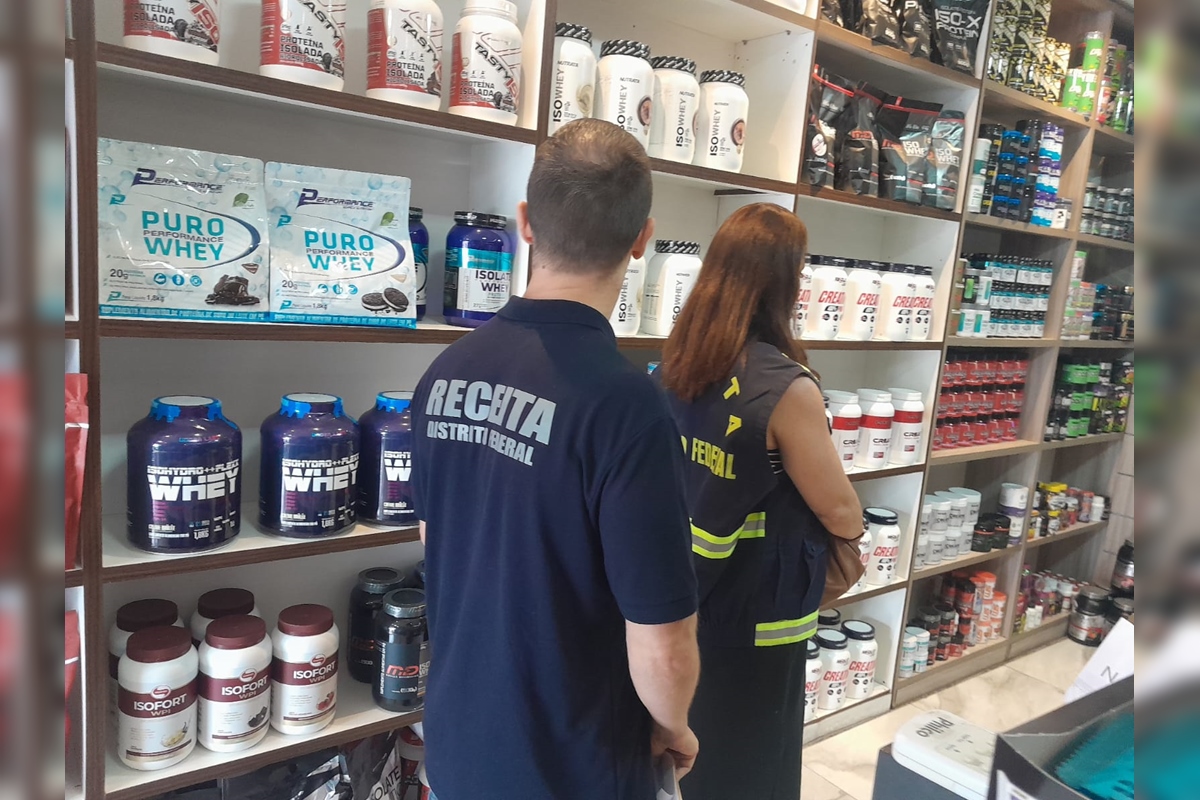 Imagem colorida de dois auditores fiscais da Receita do DF, de costas, com camisetas escrito 'Receita Distrito Federal', no interior de uma loja de suplementos alimentares. Há várias embalagens de suplementos à esquerda na imagem