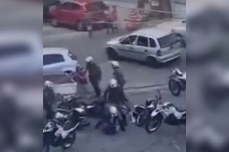 Imagem colorida mostra 5 motos paradas e 5 policiais fardados com capacetes brancos. Dois deles estão sobre um homem deitado no chão e outro PM segura uma mulher - Metrópoles