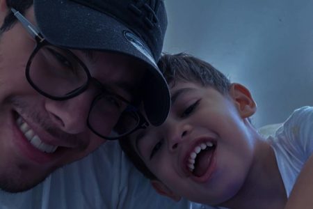 foto colorida e escura de rosto de homem de oculos e boné e rosto de menino pequeno, os dois sorriem - metrópoles