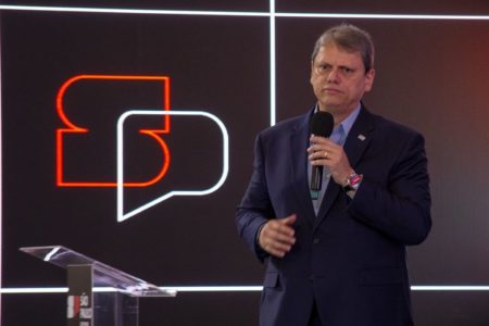 Imagem colorida mostra homem branco vestindo terno azul escuro e camisa azul clara. Ele está de pé e segura um microfone na mão esquerda. Ao seu lado, no palco, está um logotipo escrito SP - Metrópoles