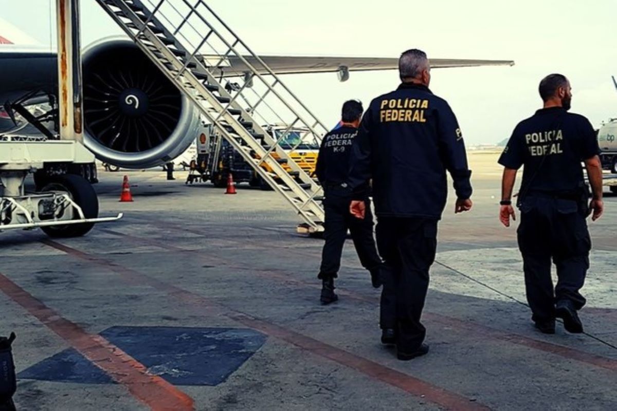 Imagem colorida mostra três homens de costas, caminhando no pátio de um aeroporto onde um avião está estacionado. Eles vestem roupas pretas com "Polícia Federal" escrito nas costas - Metrópoles