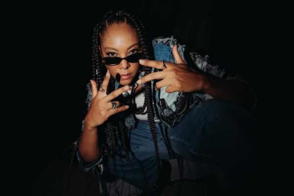 A imagem mostra a cantora Negra Li ajoelhada vestindo um conjunto de calça e jaqueta jeans