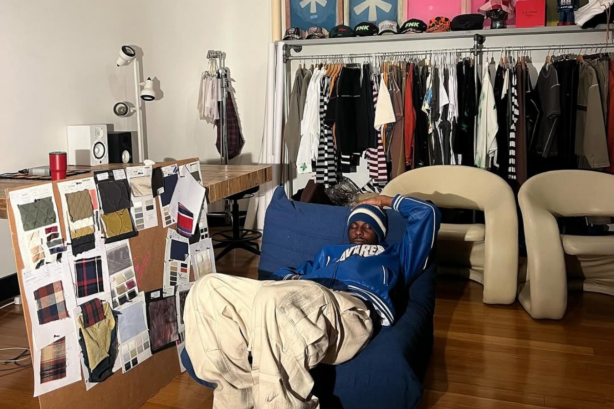 A imagem mostra um homem deitado em um sofá com diversas roupas ao fundo em um arara
