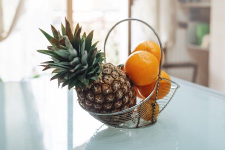 Cesta de metal com frutas - abacaxi e laranjas na mesa de vidro na cozinha. Metrópoles