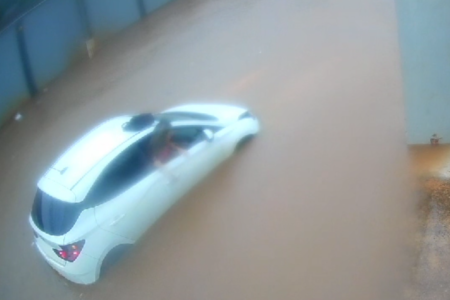 Imagem colorida de uma mulher loira, vestida com blusa rosa, descendo de um carro branco pela janela. O carro está em meio a uma enxurrada