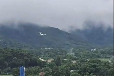 avião plana entre nuvens cinzentas e chuvas; árvores e casas compõem o plano de fundo - Metrópoles