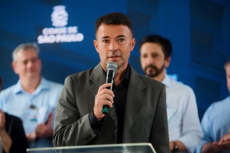 Imagem colorida mostra o vice-prefeito de SP, Coronel Mello Araújo (PL), discursando em um púlpito. Ele é um homem branco, sem barba, cabelos curtos e escuros, vestindo um paletó cinza escuro por cima de uma camisa preta e segura um microfone. Atrás há um painel da Prefeitura e também o prefeito Ricardo Nunes.