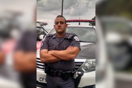 imagem colorida mostra policial militar com braços cruzados em frente à viatura da pm. ele é um homem branco, tem cabelo bem curto, e usa farda