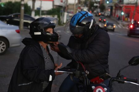 Imagem colorida mostra motociclista colocando capacete em passageira - Metrópoles