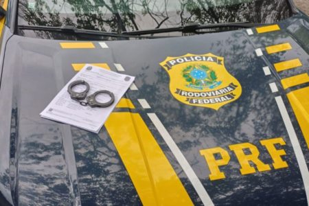 Imagens coloridas mostram capô de viatura da Polícia Rodoviária Federal com um par de algemas sobre um papel branco