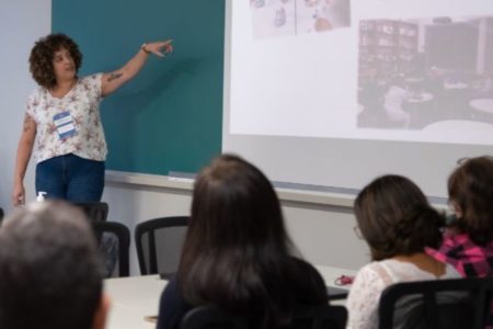 Imagem colorida mostra, no canto esquerdo e ao fundo, uma mulher com cabelos cacheados, calça jeans e camiseta clara, em frente ao quadro de uma sala de aula. Ela aponta para para uma tela. Em primeiro plano aparecem pessoas adultas sentadas de costas para a foto - Metrópoles
