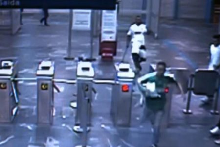 Imagem mostra print de vídeo de câmera de segurança de estação de trem em São Paulo. É possível ver catracas , um homem correndo na frente de outro homem que segura uma barra em uma das mãos - Metrópoles