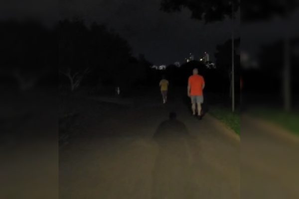 Imagem colorida de uma ciclovia de um parque, com duas pessoas caminhando. O local está escuro devido à falta de iluminação