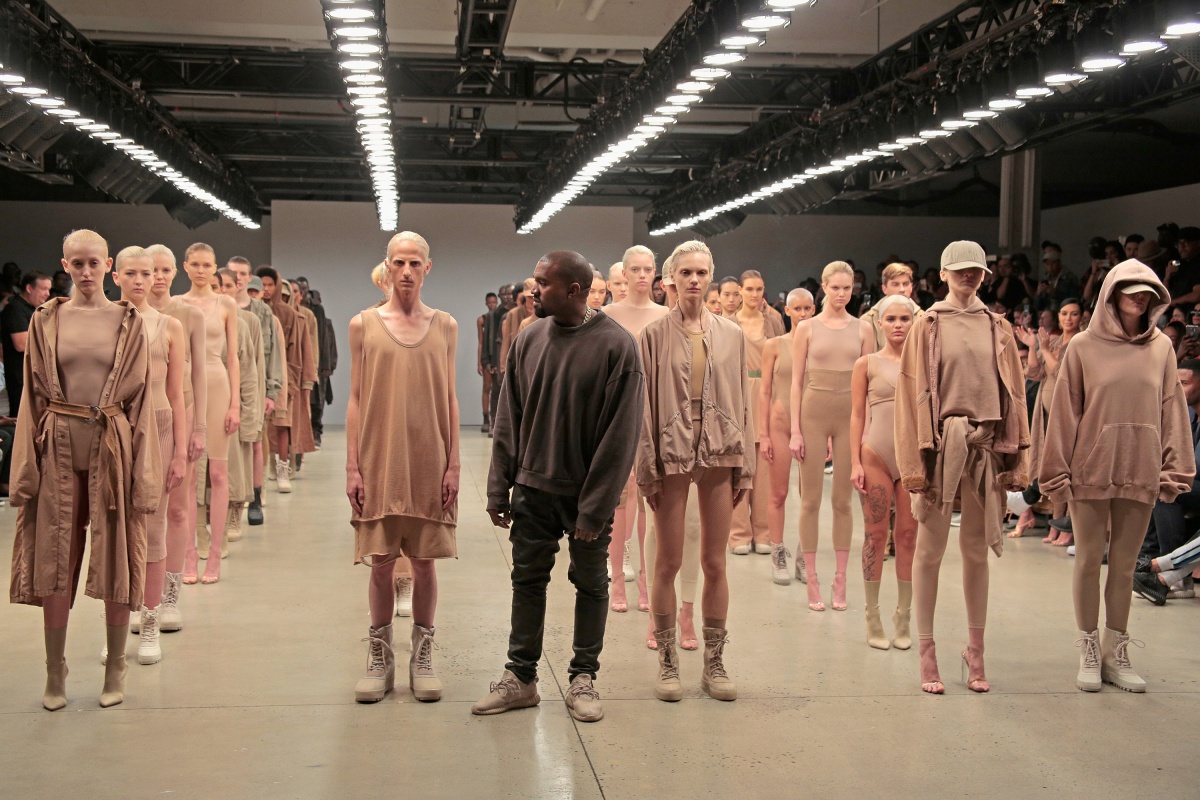 A imagem em cores mostra o artista Kanye west em frente diversos modelos vestidos com roupas em tons neutros