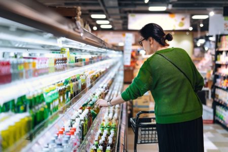 Jovem asiática com carrinho de compras pegando uma garrafa de bebida nas prateleiras refrigeradas do supermercado