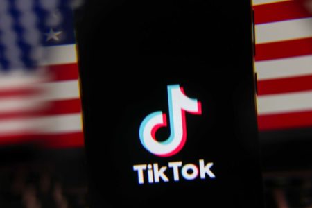 Imagem de um celular exibindo o logotipo do app TikTok e, ao fundo, uma bandeira dos Estados Unidos - Metrópoles