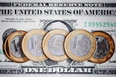 Imagem de moedas de real colocadas sobre uma nota de dólar - Metrópoles