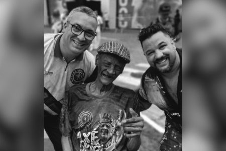 Imagem em preto e branco mostra Seu Carlão, fundador da escola de samba Unidos do Peruche, entre dois integrantes da escola - Metrópoes