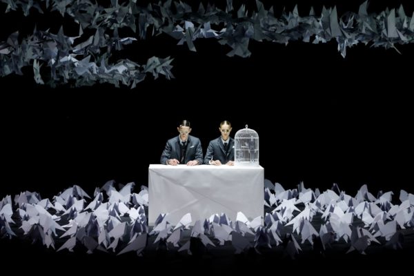 A imagem mostra dois homens sentados sob uma mesa rodeados de origamis