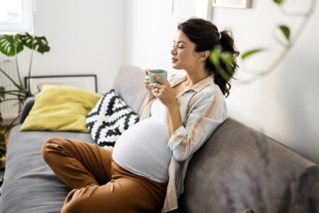 Mulher grávida descansando no sofá segurando uma xícara de café ou chá. Metrópoles