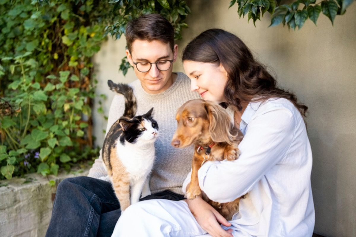 Imagem colorida de casal segurando um gato e um cachorro no colo