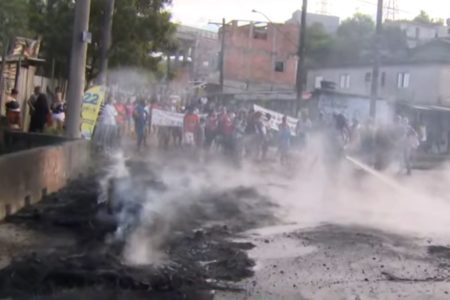 Imagem colorida mostra borracha preta, de pneus queimados, e, ao fundo, grupo de pessoas em protesto contra reintegração de posse. Elas carregam cartazes nas mãos - Metrópoles