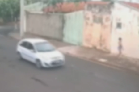 Imagem colorida de carro branco passando pela rua enquanto uma adolescente anda na calçada. Metrópoles
