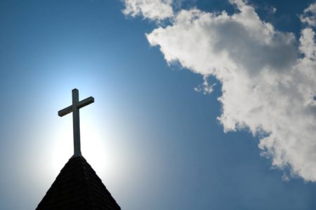 Imagem colorida de cruz em topo de igreja, aparece céu azul com poucas nuvens ao fundo - Metrópoles