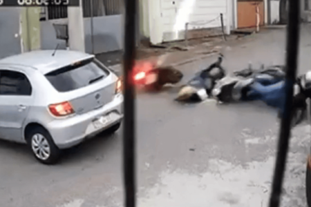 Imagem colorida de uma câmera de segurança mostra três homens caindo de motocicleta em uma via. Além disso, um deles está em rota de colisão com um carro prata parado. Um motociclista alvo de tentativa de assalto foi levado para o hospital - Metrópoles