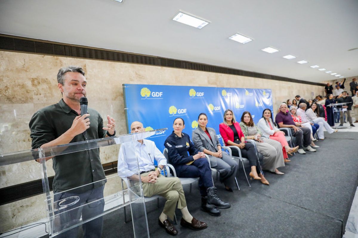 Imagem colorida, com o secretário Gustavo Rocha em pé com o microfone, em um palco com autoridades sentadas