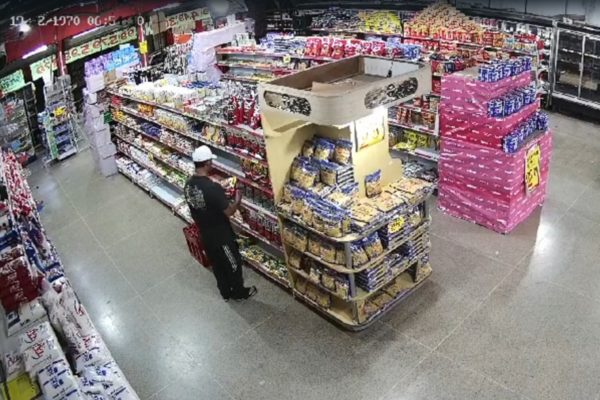 Imagem colorida de um homem de camiseta preta, calça preta e boné branco escolhendo itens em um supermercado