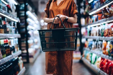 Foto recortada de uma jovem carregando uma cesta de compras, em pé ao longo do corredor de produtos, fazendo compras de necessidades diárias no supermercado. Metrópoles