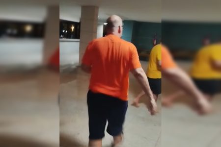 Imagem colorida de homem careca com camisa laranja em pilotis de prédio