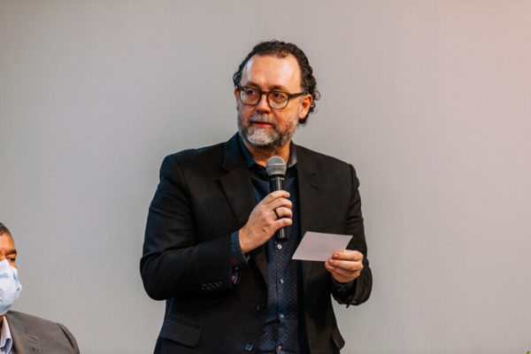 Fotografia colorida mostra Carlos Bezerra Júnior, da cintura para cima, um homem de barbas grisalhas, óculos redondo, vestindo terno preto e segurando um microfone com a mão direita e um papel com a esquerda - Metrópoles