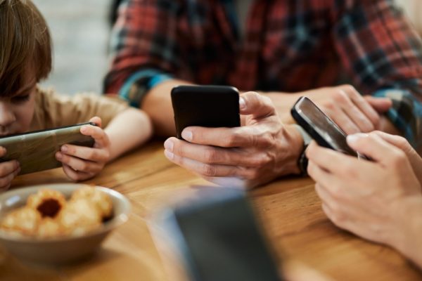 Foto colorida de pais e crianças sentados em uma mesa mexendo no celular - Metrópoles