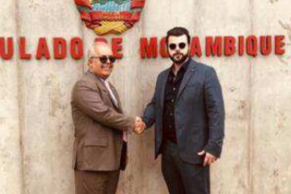 Dois homens vestidos com ternos apertam as mãos, em frente a muro com os dizeres 