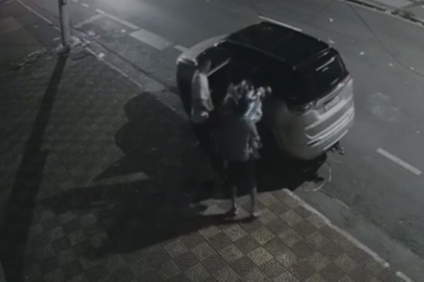 Imagem em preto e branco de dois homens com sacolas de lixo ao lado de carro de luxo. Metrópoles
