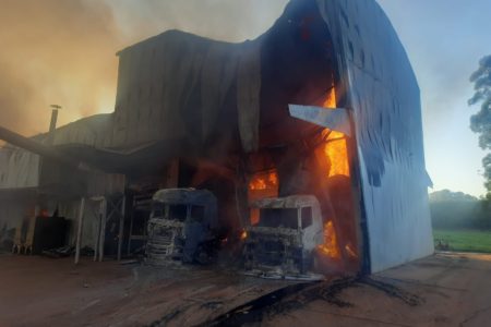 Imagem colorida de caminhões pegando fogo dentro de galpão em chamas. Metrópoles