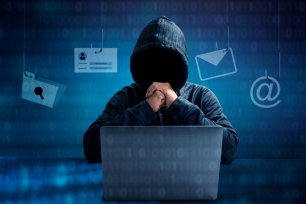Imagem de homem com capuz, rosto encoberto, sentado em frente a um notebook - Metrópoles