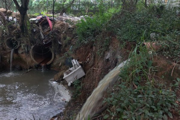imagem colorida mostra líquido amarelado saindo de tubulação e caindo em lugar de mata, onde há restos de móveis, lixo e outras tubulações