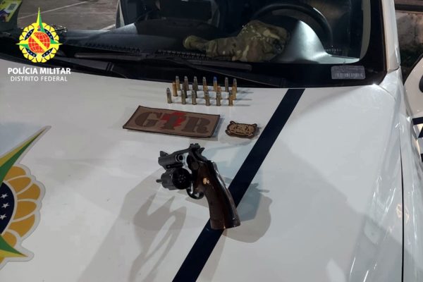 Imagem colorida de uma arma e diversas munições sobre uma viatura branca com detalhes azuis da PMDF