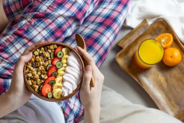 Imagem colorida de pessoa sentada na cama, comendo frutas
